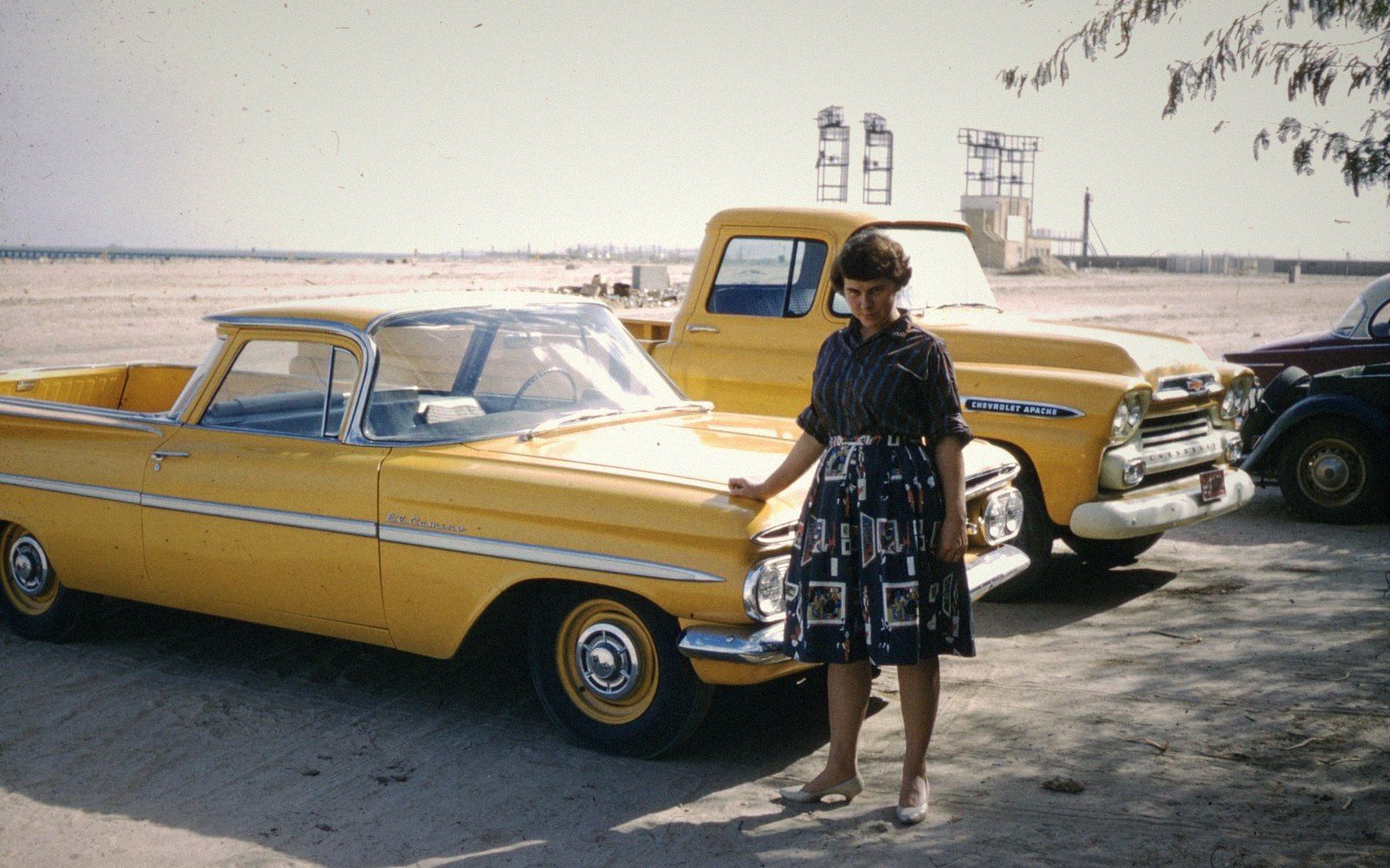 A woman in a patterned dress stands beside vintage yellow Chevrolet trucks on a sandy lot